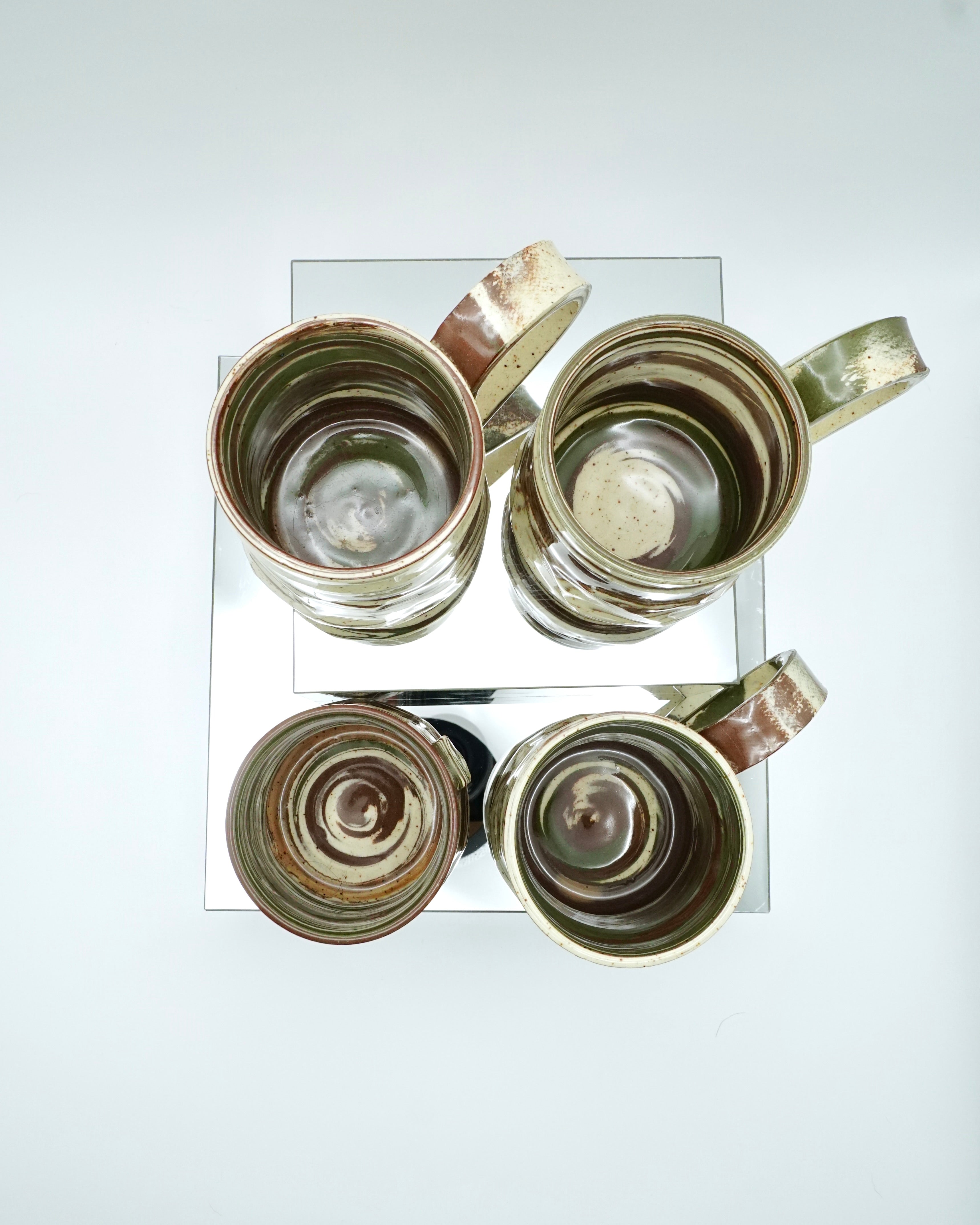 Set of four ceramic mugs with brown, green, and speckled white abstract patterns on reflective surfaces and a white background.