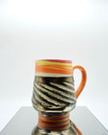 Ceramic mug with red, orange, yellow, white swirled patterns and chrome glaze on a reflective surface and white background.