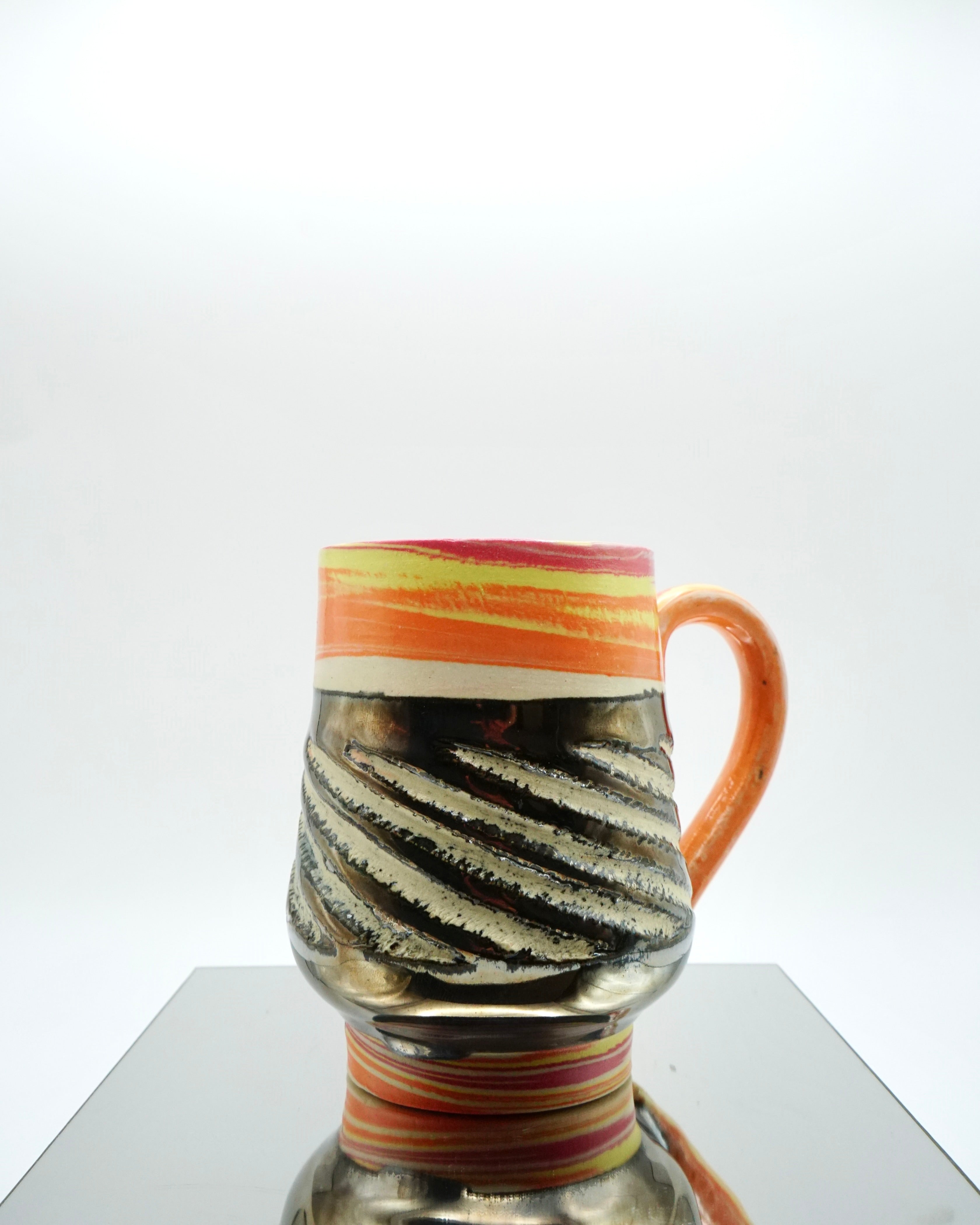 Ceramic mug with red, orange, yellow, white swirled patterns and chrome glaze on a reflective surface and white background.