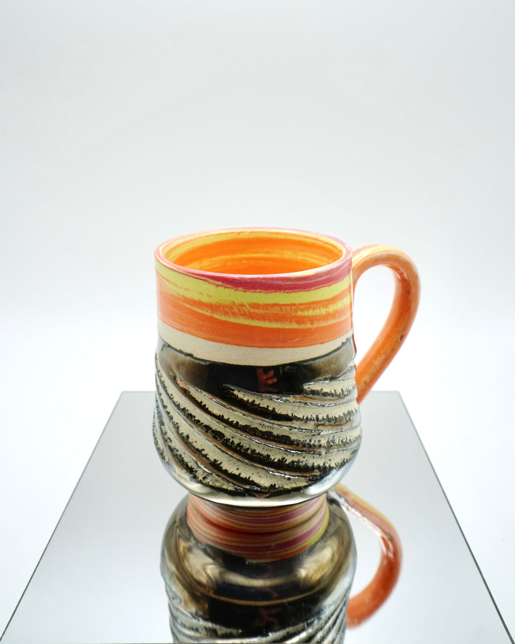 Ceramic mug with red, orange, yellow, white swirled patterns and chrome glaze on a reflective surface and white background.