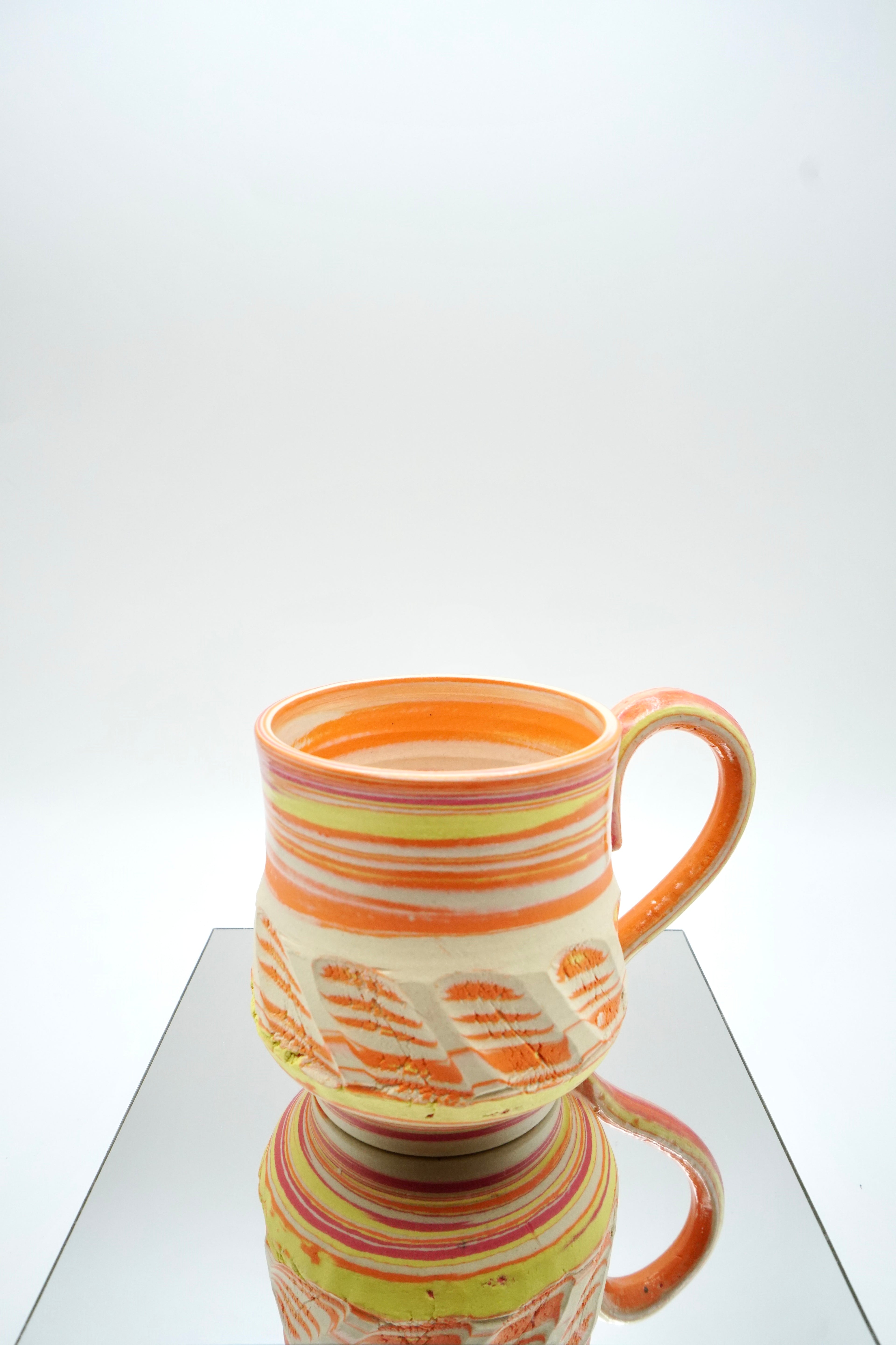 Ceramic cup with red, orange, yellow, white swirled patterns on a reflective surface and white background.