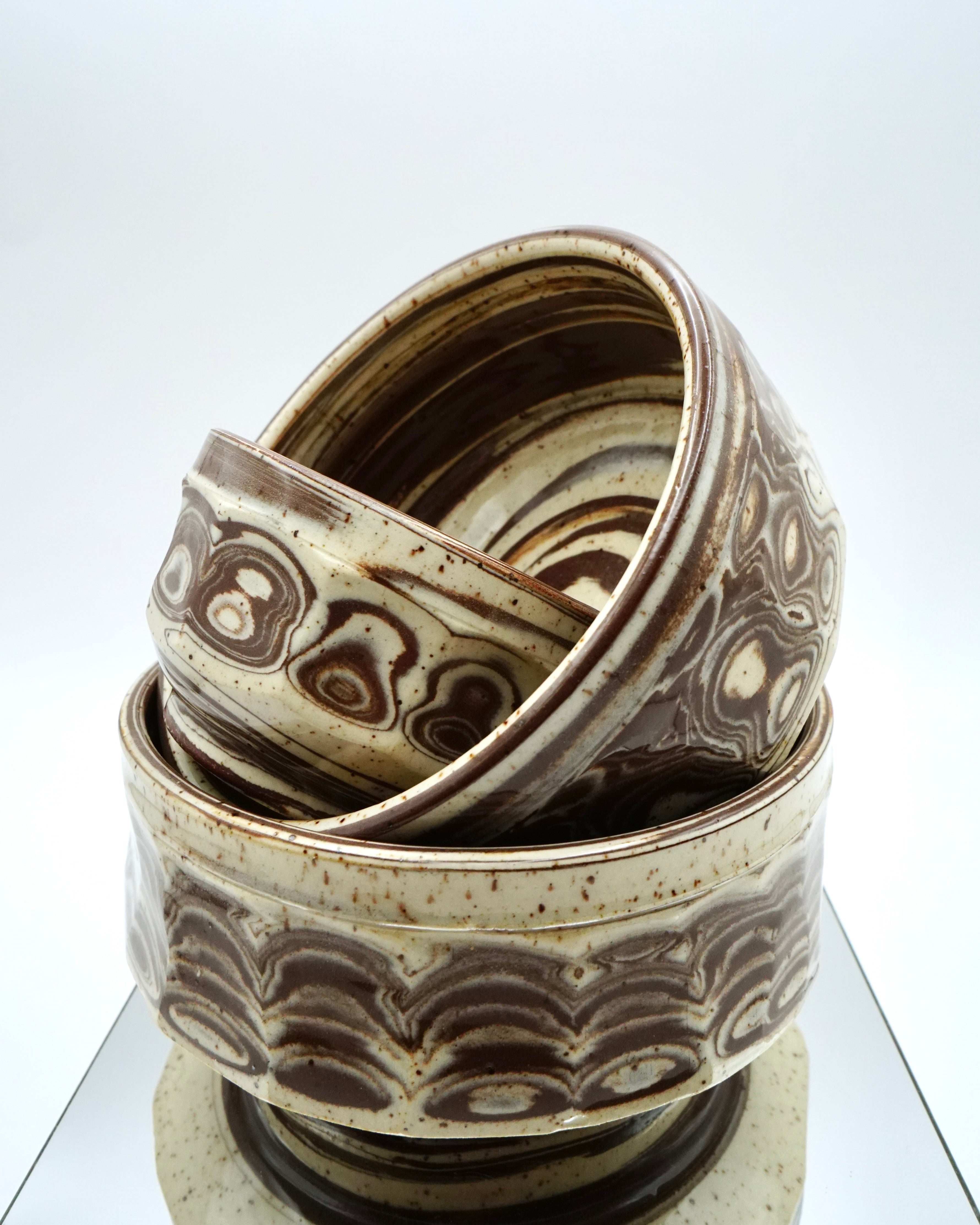 Stack of ceramic bowls with brown and speckled white patterns on a reflective surface and white background.