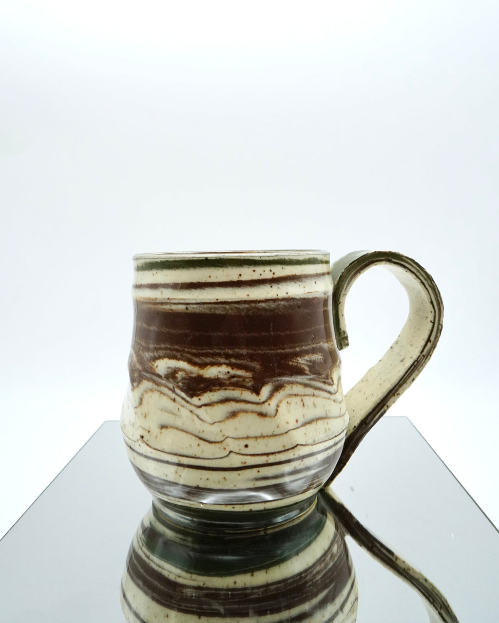 Ceramic mug with brown, green, and speckled white abstract patterns on a reflective surface and white background.