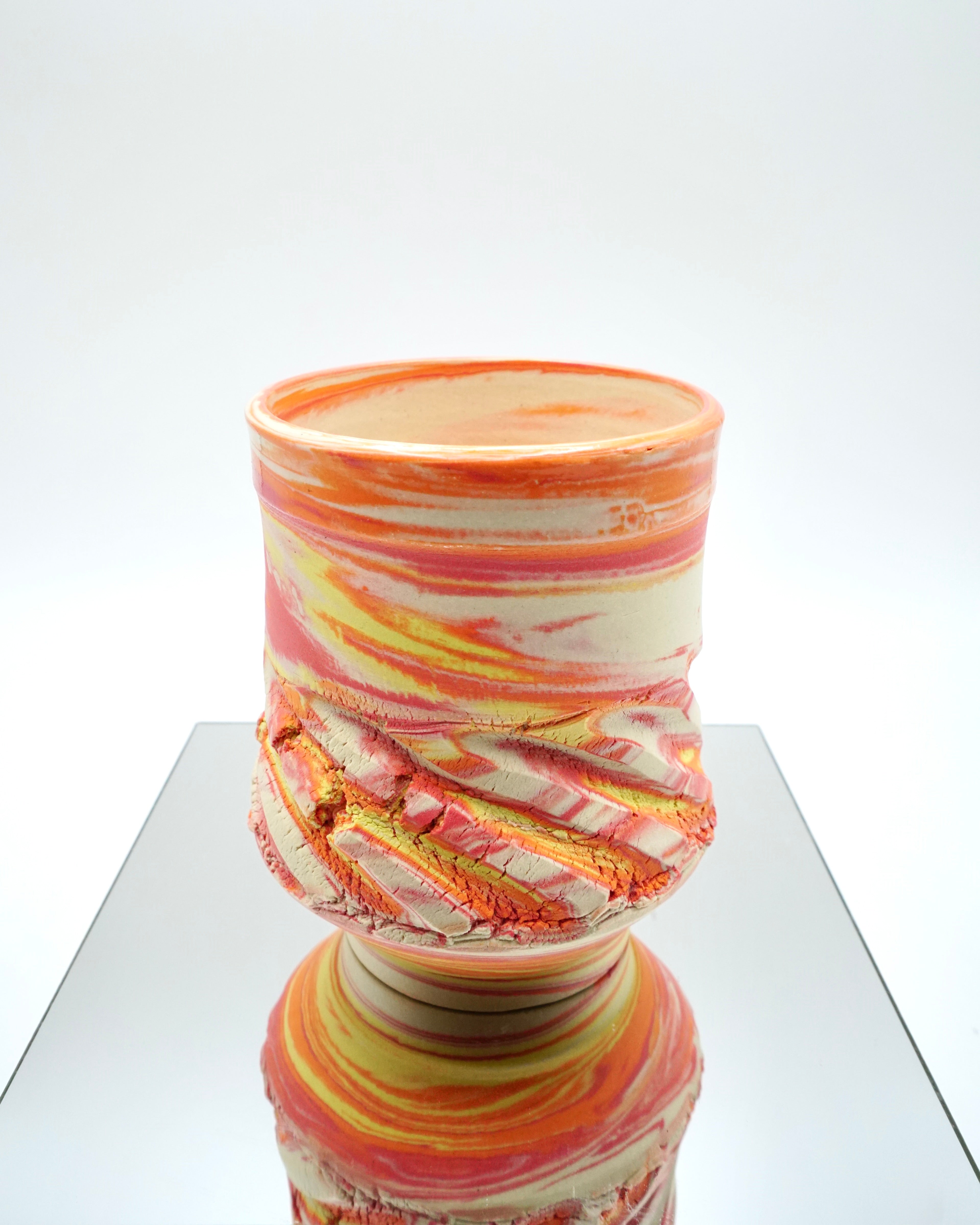 Ceramic cup with red, orange, yellow, white swirled patterns on a reflective surface and white background.