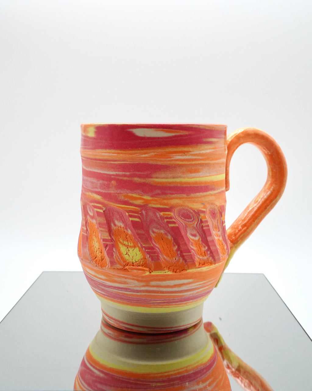 Ceramic mug with red, orange, yellow, white swirled patterns on a reflective surface and white background.